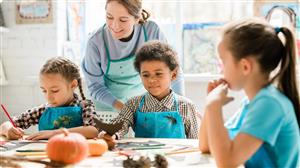 elementary kids in apron working at a table; teacher in apron guiding them